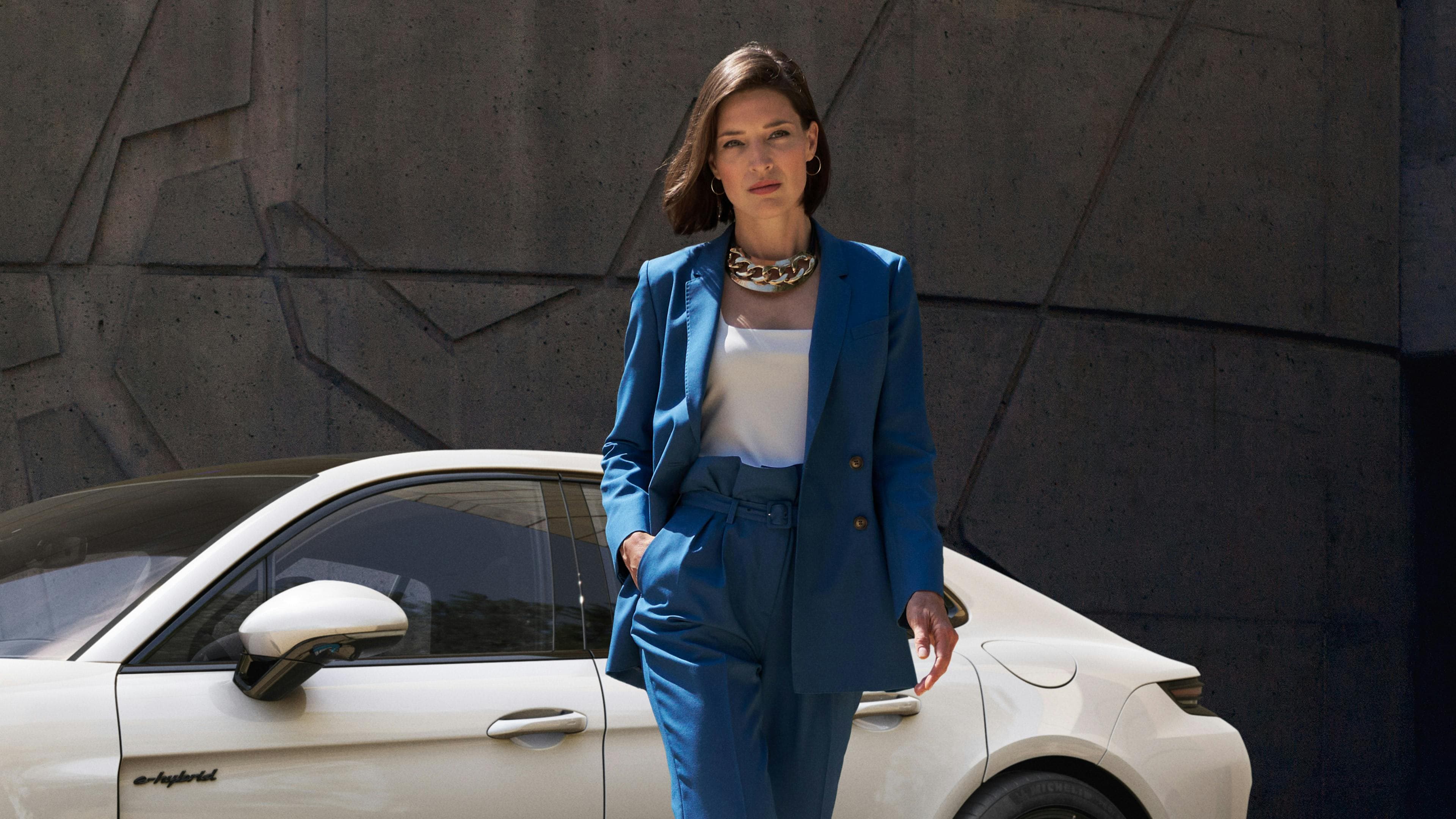 A woman in a blue suit stands confidently next to a white Porsche against a textured concrete wall.