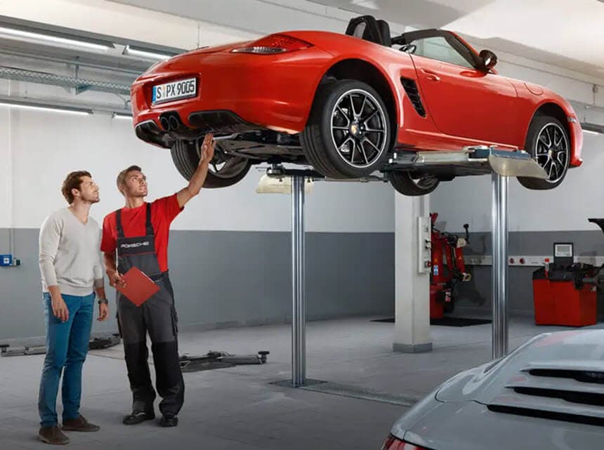 Two men inspect a red convertible car lifted on a garage hoist. One man points underneath, while the other observes.